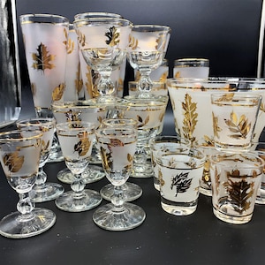 May include: A collection of frosted glassware featuring gold leaf patterns, including various stemmed glasses, tumblers, and a large serving bowl. Each piece is decorated with metallic gold foliage designs and thin gold rims on clear glass bases.