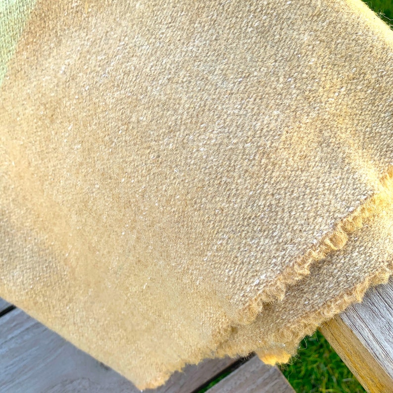 May include: A close-up of a light brown woven blanket with a fringed edge. The blanket is folded and draped over a wooden surface.