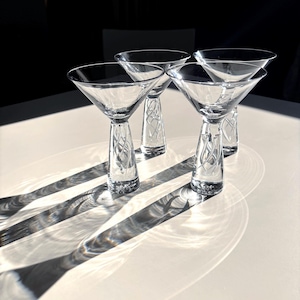 May include: Four clear glass martini glasses with a decorative stem design. The glasses are arranged on a white surface, casting long shadows in the sunlight. The stems have an etched, swirling pattern. The glasses are empty.