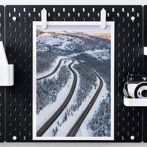 Puede incluir: Una fotografía en blanco y negro de un camino sinuoso a través de un paisaje montañoso nevado. El camino es visible en el centro de la imagen, con árboles y montañas cubiertos de nieve a ambos lados.