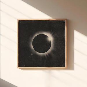 May include: A framed print of a solar eclipse. The artwork shows a dark, textured background with a central black circle representing the moon, encircled by a bright, radiant corona. The frame is made of light-coloured wood.