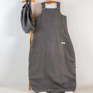 Könnte beinhalten: Ein grauer Waffelstoff-Schlafsack für Babys mit weißen Trägern und einem weißen Etikett. Der Schlafsack hängt an einer Holzleiter, über ihm hängt eine graue Waffelstoff-Tasche.