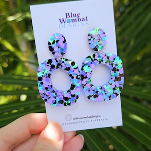 May include: A pair of iridescent purple and green statement earrings with a circle cutout. The earrings are made of resin and have a confetti-like design. The earrings are handcrafted in Australia. @bluewombatdesigns