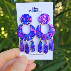 May include: Glittery purple earrings with a circular top, a crescent-shaped middle, and teardrop pendants. The earrings feature blue and pink glitter. The brand name "Blue Wombat Designs" is visible. Handcrafted in Australia.