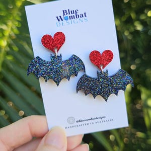 May include: A pair of dangle earrings featuring a red glitter heart attached to a black glitter bat shape. The earrings are displayed on a white card with the text Blue Wombat DESIGNS and the social media handle @bluewombatdesigns printed on it.