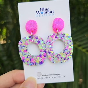May include: A pair of handmade, glitter earrings. Each earring has a bright pink, oval top and a square bottom with a rounded hole. The earrings are filled with colourful glitter and are attached to a white card with the text "Blue Wombat Designs". Handcrafted in Australia.
