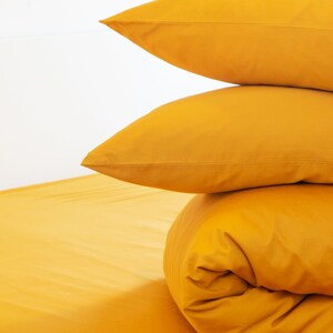 May include: A stack of three golden-yellow bed linens against a white background. Two pillowcases are stacked on top of a rolled-up comforter, with a sheet visible at the bottom. The bedding is a solid color.