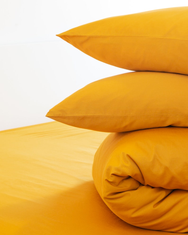 May include: A stack of three golden-yellow bed linens against a white background. Two pillowcases are stacked on top of a rolled-up comforter, with a sheet visible at the bottom. The bedding is a solid color.
