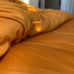 May include: A soft, golden-yellow duvet cover draped over a bed, featuring gentle folds and creases. The fabric surface is illuminated by warm, directional sunlight, creating subtle highlights and shadows across the textured material.