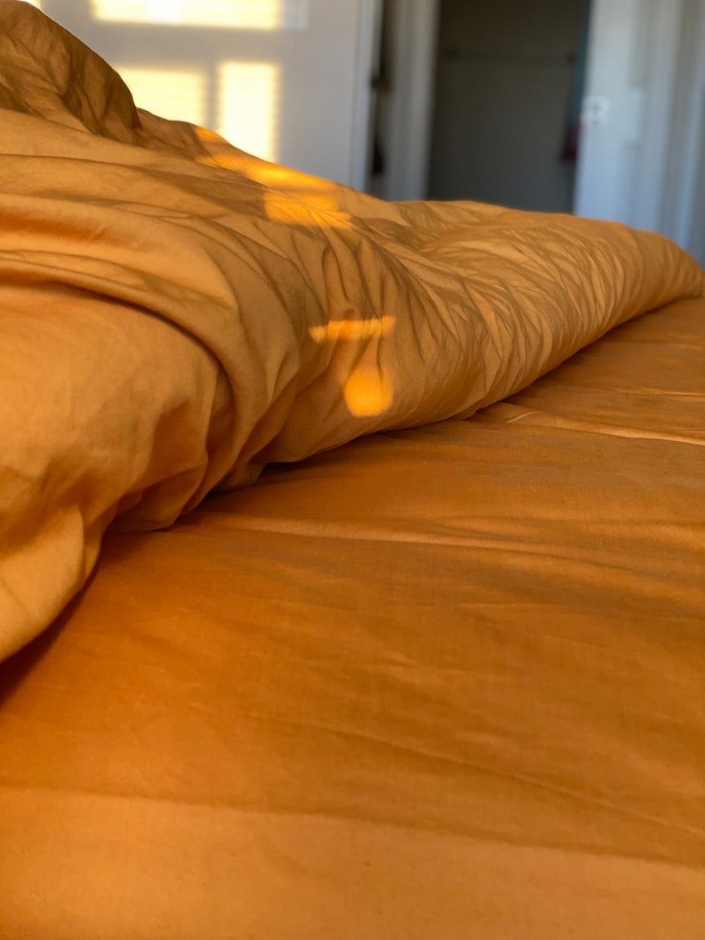 May include: A soft, golden-yellow duvet cover draped over a bed, featuring gentle folds and creases. The fabric surface is illuminated by warm, directional sunlight, creating subtle highlights and shadows across the textured material.