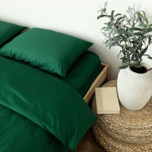 Op de afbeelding: Een bed met smaragdgroen beddengoed, inclusief een hoeslaken en twee kussenslopen. Een witte vaas met groen staat op een geweven kruk naast een klein boek. Het bedframe is gemaakt van lichtgekleurd hout.