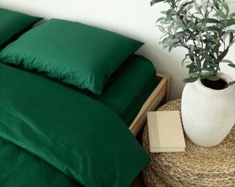 May include: A bed with emerald green bedding, including a fitted sheet and two pillowcases. A white vase with greenery sits on a woven stool next to a small book. The bed frame is made of light-colored wood.