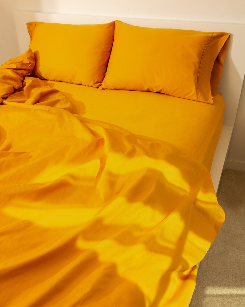May include: A bed with bright yellow bedding. Two matching pillowcases rest on the bed, with a fitted sheet and a top sheet. The sheets are slightly rumpled, and the bed frame is white.
