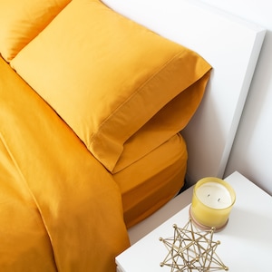 May include: A mustard yellow bedding set featuring a flat sheet and a pillowcase. Next to the bed is a white nightstand holding a yellow glass candle with two wicks and a gold-toned geometric metal sculpture made of connected rods.