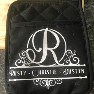 Może przedstawiać: Czarna pikowana rękawica kuchenna z białym dekoracyjnym wzorem i imionami "Rusty - Christie - Dustyn". Wzór zawiera dużą literę "R" w okręgu z dekoracyjnymi zawijasami.