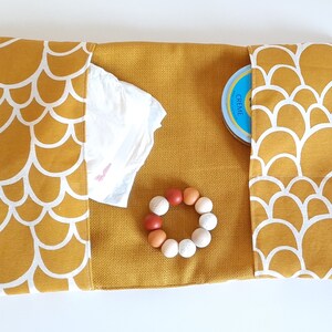 May include: A mustard yellow changing mat with white scalloped edges. The mat has two pockets, one containing a blue container with the word "CREME" on it, and the other containing a white diaper. A wooden teething ring with white and orange beads is in the center of the mat.