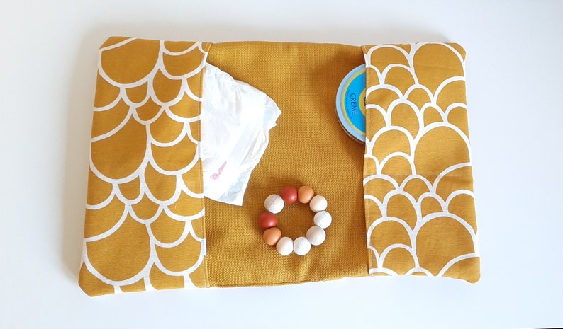 May include: A mustard yellow changing mat with white scalloped edges. The mat has two pockets, one containing a blue container with the word "CREME" on it, and the other containing a white diaper. A wooden teething ring with white and orange beads is in the center of the mat.