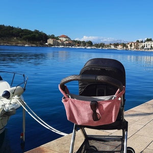 May include: A black stroller with a pink fabric storage bag hanging from the handle. The stroller is parked on a concrete walkway near a body of water. Sunglasses are in the storage bag.