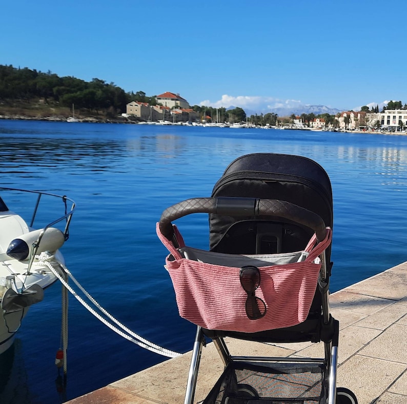 May include: A black stroller with a pink fabric storage bag hanging from the handle. The stroller is parked on a concrete walkway near a body of water. Sunglasses are in the storage bag.