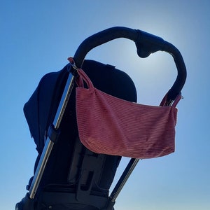 May include: A black stroller with a pink corduroy bag attached to the handle. The stroller is against a bright blue sky.