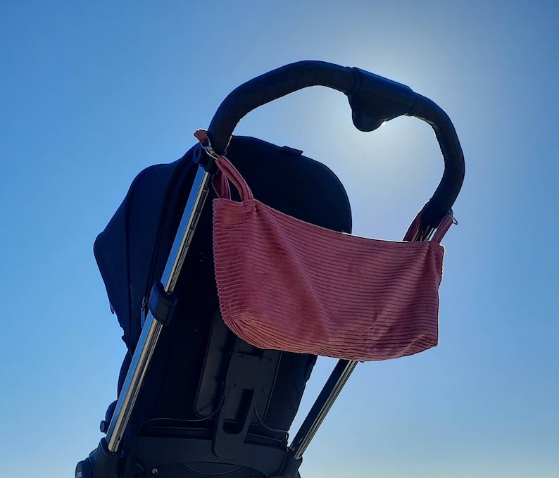 May include: A black stroller with a pink corduroy bag attached to the handle. The stroller is against a bright blue sky.