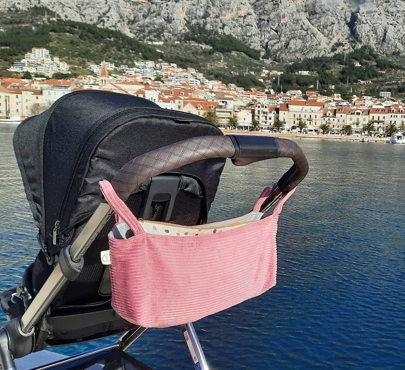 May include: A pink stroller organizer with a white and brown patterned lining hanging on a black stroller. The stroller is parked in front of a scenic view of a city and a body of water.