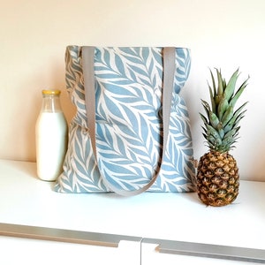 May include: A blue and white tote bag with a leaf pattern and gray straps. The bag is sitting on a white surface with a pineapple and a bottle of milk.