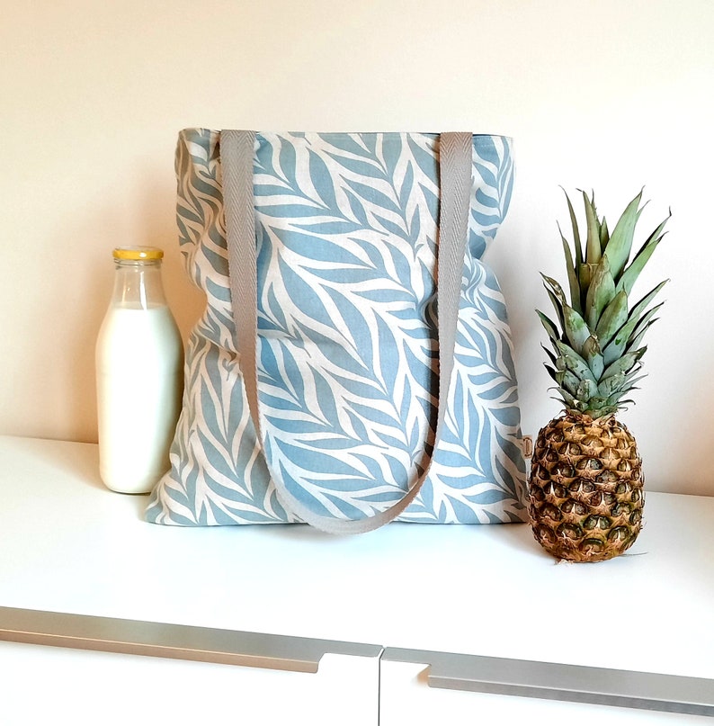 May include: A blue and white tote bag with a leaf pattern and gray straps. The bag is sitting on a white surface with a pineapple and a bottle of milk.
