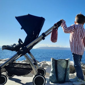 May include: A black stroller with a gray and white striped bag hanging from the handle. A child is standing next to the stroller, wearing a red and white striped shirt and blue jeans.