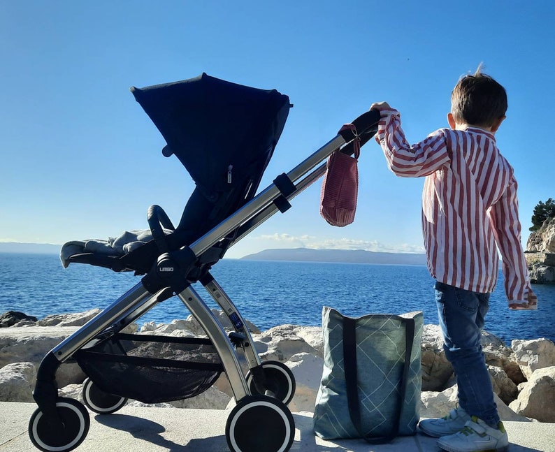 May include: A black stroller with a gray and white striped bag hanging from the handle. A child is standing next to the stroller, wearing a red and white striped shirt and blue jeans.