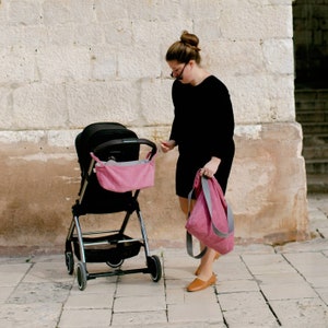 May include: A woman in a black dress pushes a black stroller with a pink fabric liner. She is carrying a pink tote bag with a gray strap.