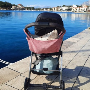 May include: A black stroller with a pink and white polka dot bag attached to the handle. The stroller is on a stone walkway with a body of water in the background.