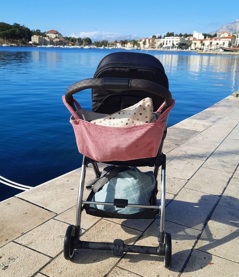 May include: A black stroller with a pink and white polka dot bag attached to the handle. The stroller is on a stone walkway with a body of water in the background.