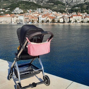 May include: A black stroller with a pink storage bag, parked on a concrete surface near a body of water. The stroller has a black fabric seat and a silver frame. The storage bag is attached to the stroller's handle.