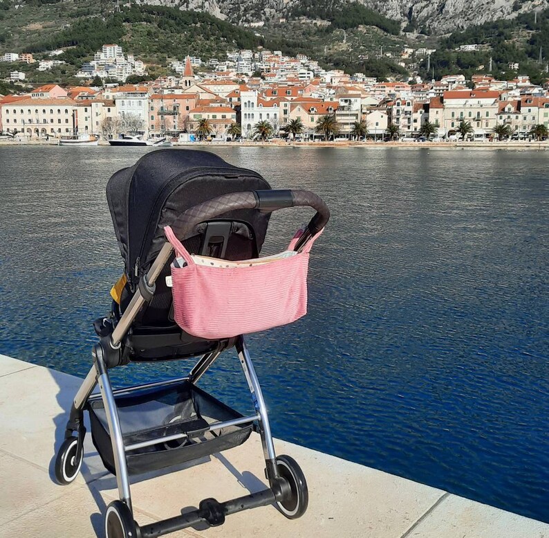 May include: A black stroller with a pink storage bag, parked on a concrete surface near a body of water. The stroller has a black fabric seat and a silver frame. The storage bag is attached to the stroller's handle.