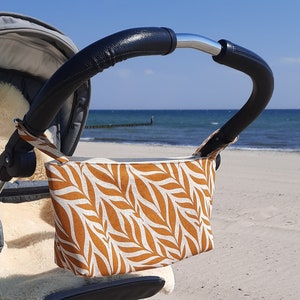 May include: A brown and white patterned fabric stroller organizer hanging on a stroller handle. The organizer is made of a soft, durable fabric and has a zipper closure. The stroller is parked on a sandy beach.