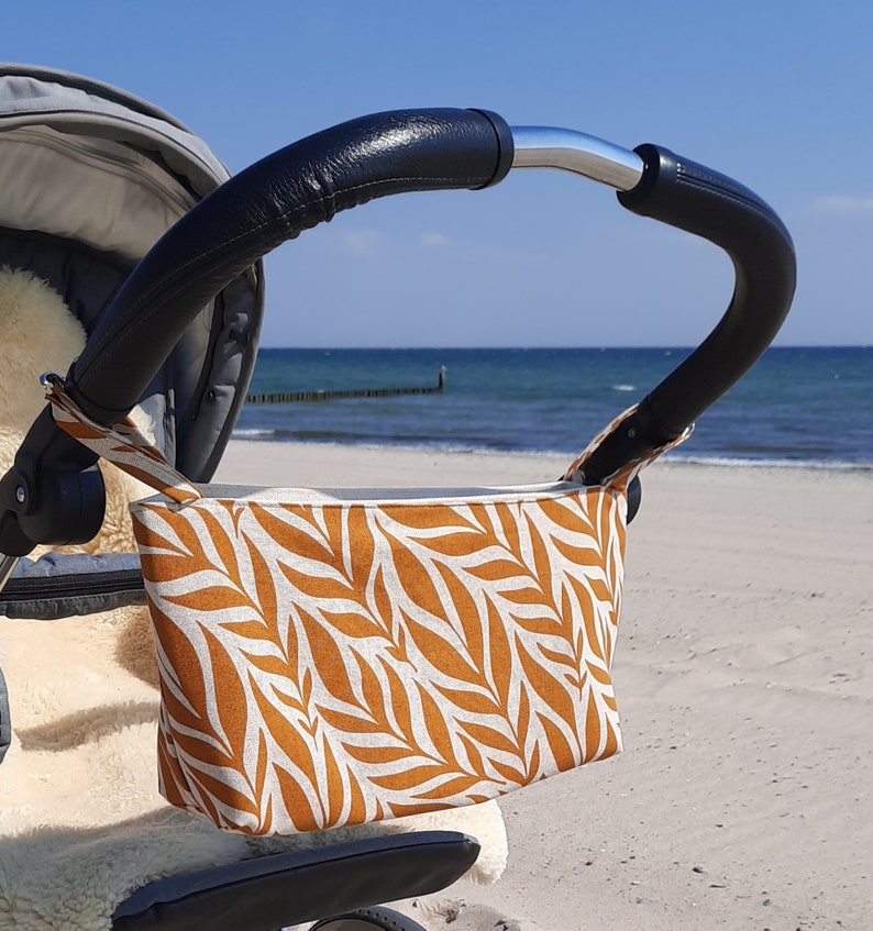 May include: A brown and white patterned fabric stroller organizer hanging on a stroller handle. The organizer is made of a soft, durable fabric and has a zipper closure. The stroller is parked on a sandy beach.