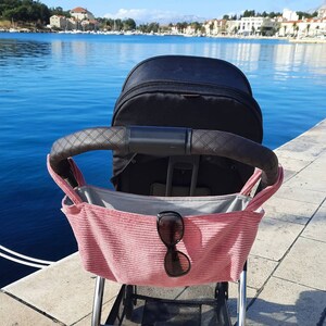 May include: A black stroller with a pink fabric storage bag hanging from the handle. The bag has a pair of sunglasses inside. The stroller is parked on a stone walkway overlooking a body of water.