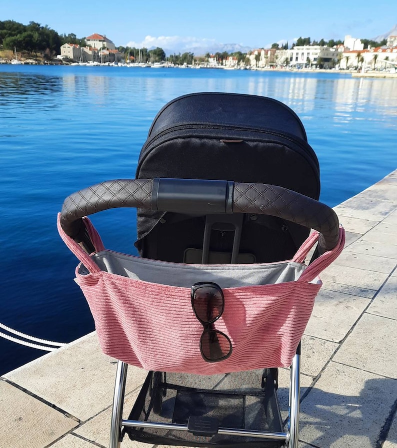 May include: A black stroller with a pink fabric storage bag hanging from the handle. The bag has a pair of sunglasses inside. The stroller is parked on a stone walkway overlooking a body of water.