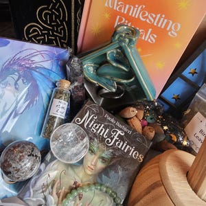 May include: A collection of spiritual items including a book titled "Manifesting Rituals", a teal ceramic sculpture, a crystal ball, a deck of oracle cards, and a bottle of herbs. The items are arranged in a wooden box.