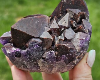 May include: A cluster of dark, reddish-brown crystals with deep purple amethyst formations at the base. The specimen features sharp, geometric points and a rough, textured exterior, showcasing a natural mineral structure with varied dark tones.