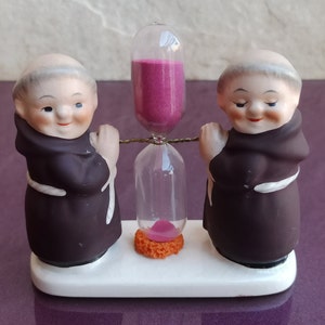 Può includere: Due figurine in ceramica di monaci in abiti marroni che tengono una clessidra rosa con dettagli dorati. La clessidra è su una base bianca.