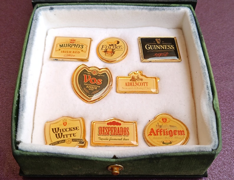 May include: A collection of eight beer bottle cap pins in a green velvet box. The pins feature logos for various beer brands, including Murphys Irish Red, Fischer, Guinness, Vos, Adelscott, Wieckse Witte, Desperados, and Affligem.