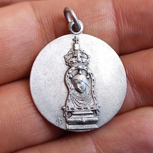 May include: A silver-colored pendant with a detailed relief of a crowned figure within an arched frame. The pendant has a circular shape with a loop at the top for a chain. The design includes intricate details.