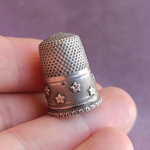 May include: A silver thimble with a textured surface and a band of star-shaped cutouts. The thimble is a sewing tool, designed to protect the finger while pushing a needle through fabric. The thimble is held in a hand.