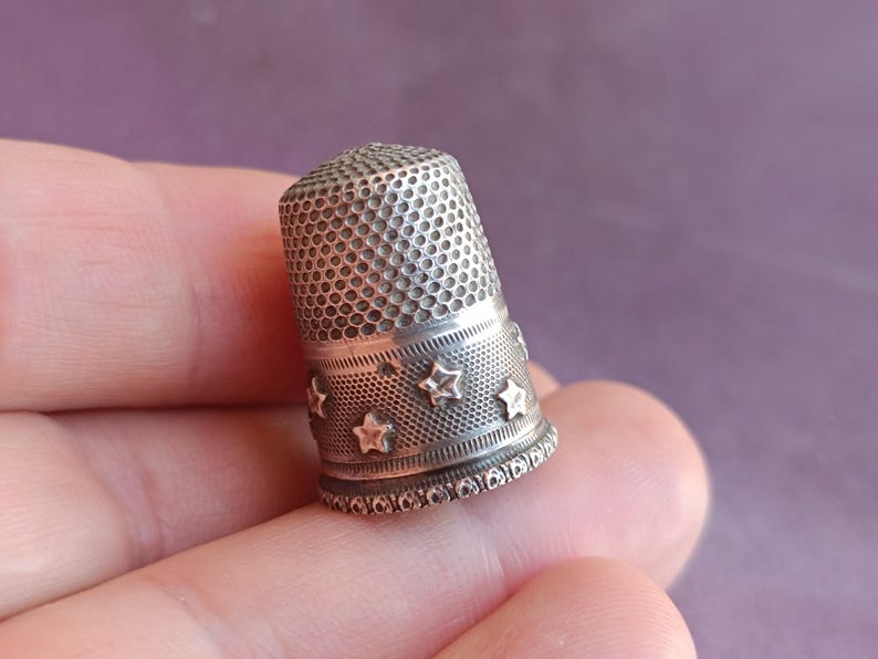 May include: A silver thimble with a textured surface and a band of star-shaped cutouts. The thimble is a sewing tool, designed to protect the finger while pushing a needle through fabric. The thimble is held in a hand.
