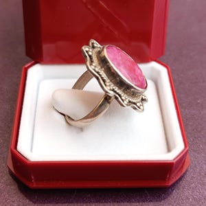 Op de afbeelding: Een zilveren ring met een grote, ovale, roze edelsteen, gezet in een decoratieve bezel. De ring wordt gepresenteerd in een rode sieradendoos met een wit interieur, die het ontwerp en de kleur benadrukt.