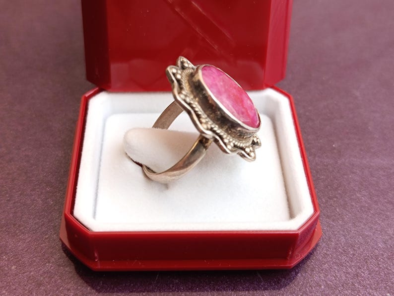 Op de afbeelding: Een zilveren ring met een grote, ovale, roze edelsteen, gezet in een decoratieve bezel. De ring wordt gepresenteerd in een rode sieradendoos met een wit interieur, die het ontwerp en de kleur benadrukt.