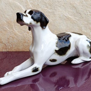 Puede incluir: Una figurita de porcelana blanca de un perro Braco Alemán de Pelo Corto blanco y negro acostado. El perro tiene ojos marrones y nariz negra.