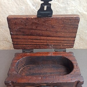May include: A small wooden box with a hinged lid and a metal latch. The box is made of dark wood and has a simple design. The inside of the box is empty.